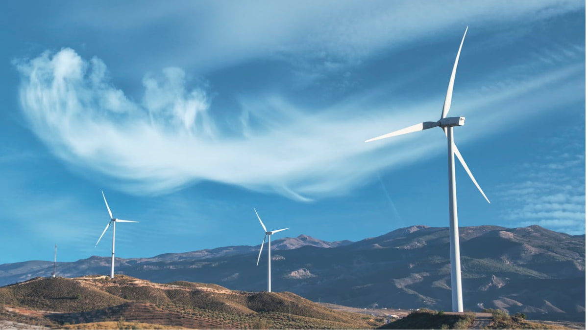 Turbinas eólicas em área montanhosa sob céu azul, representando a geração de energia limpa por fontes renováveis.