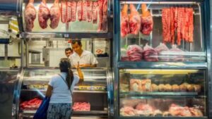 Uma cliente em frente ao balcão de um açougue, com cortes de carne pendurados e embalados à mostra.