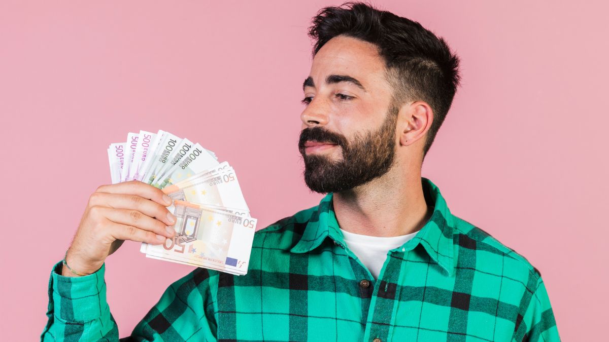 Homem com camisa quadriculada verde, segurando dinheiro na frente de uma parede rosa.