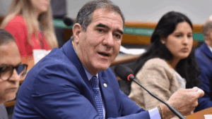 Luiz Gastão, deputado federal, discute sobre o adicional da CSLL para multinacionais durante sessão parlamentar, com outros participantes ao fundo.
