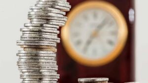 Imagem com moedas de prata uma sobre as outras e relógio ao fundo. Fim da taxa de custódia a partir de janeiro de 2025