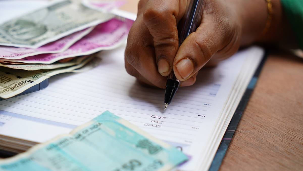 Na imagem, uma mão de uma pessoa negra faz anotações em meio a cédulas de dinheiro, um sinal da inadimplência dos consumidores