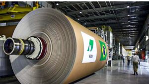 Grande rolo de papel em fábrica da Klabin com destaque para o logo da empresa e a bandeira do Brasil, representando sustentabilidade.