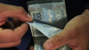 Notas de R$ 100 sobrepostas em uma mão, que as conta. Saiba qual banco deu mais lucro aos acionistas