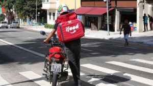 Entregador do iFood em moto atravessa rua, destacando a discussão sobre direitos trabalhistas no setor de delivery.