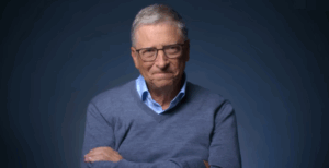 Bill Gates em um ambiente de fundo escuro, usando óculos, camisa azul clara e suéter cinza, com os braços cruzados, refletindo sobre o futuro da inteligência artificial.