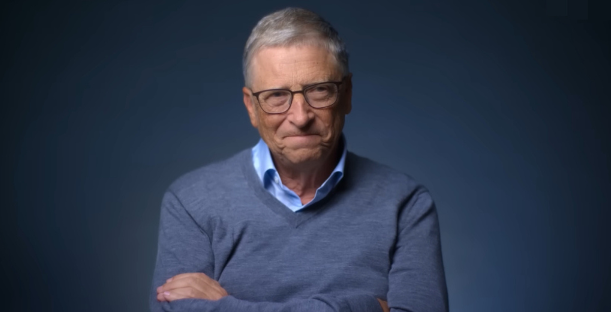Bill Gates em um ambiente de fundo escuro, usando óculos, camisa azul clara e suéter cinza, com os braços cruzados, refletindo sobre o futuro da inteligência artificial.