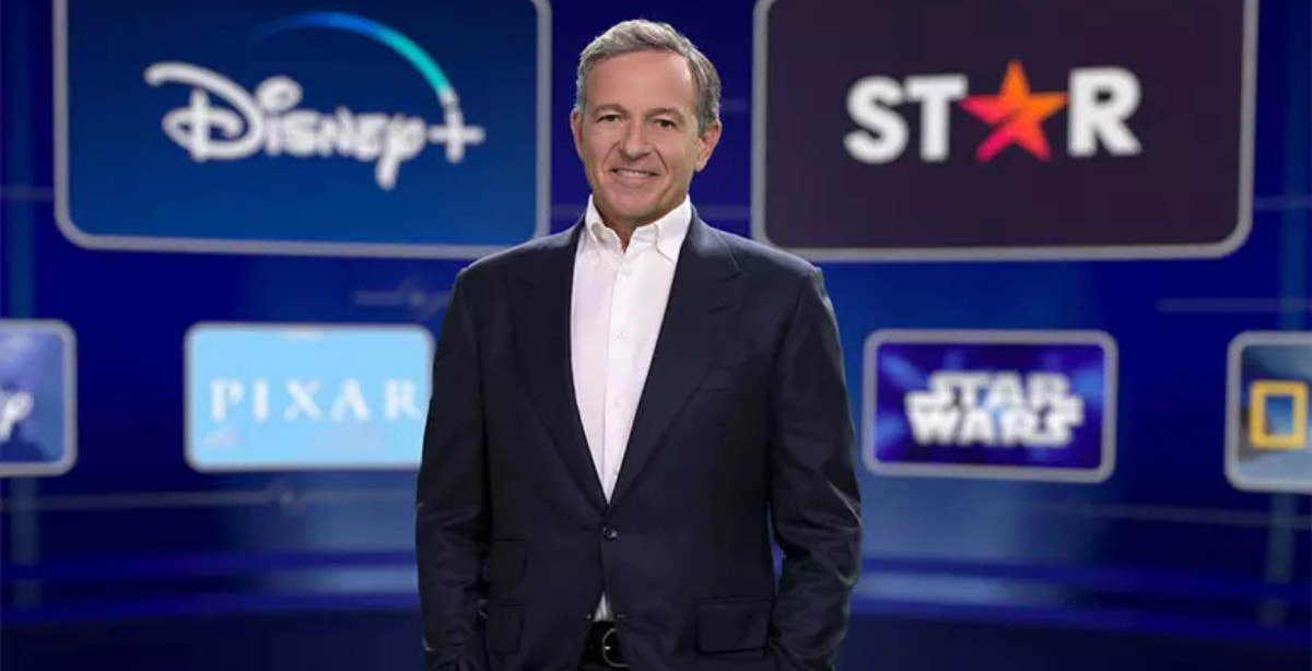 Bob Iger, CEO da Disney, em pé, sorrindo, com logotipos da Disney+, Star, Pixar, Star Wars e National Geographic ao fundo.