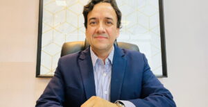 Frederico Cortez sentado em um escritório, vestindo terno azul, camisa listrada, com fundo decorativo geométrico ao fundo.