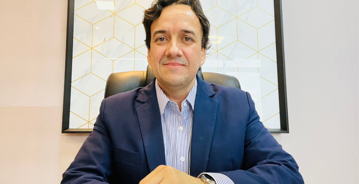 Frederico Cortez sentado em um escritório, vestindo terno azul, camisa listrada, com fundo decorativo geométrico ao fundo.