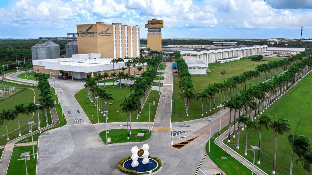 Vista aérea do complexo industrial da M. Dias Branco em expansão, destacando sua moderna estrutura com edifícios administrativos, silos de armazenamento e uma ampla área verde com palmeiras e vias bem planejadas. O cenário reflete o crescimento da empresa no setor alimentício.
