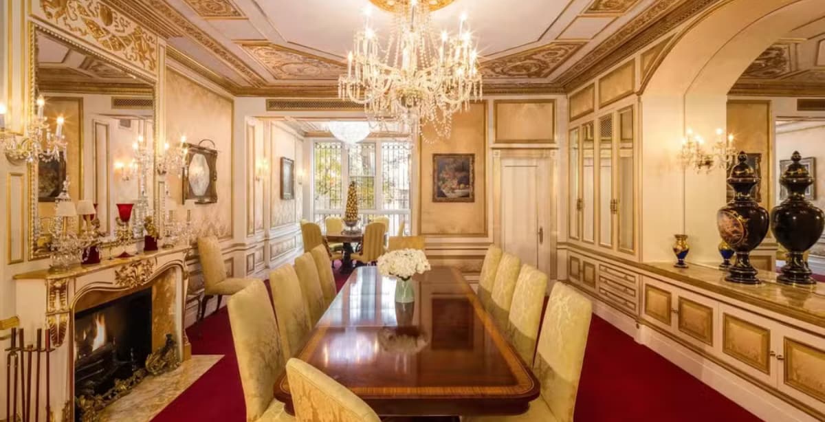 Luxuosa sala de jantar da mansão de Ivana Trump em Nova York, com decoração clássica em tons dourados, candelabros de cristal, lareira elegante e uma mesa de madeira com cadeiras estofadas.