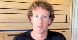Mark Zuckerberg em ambiente interno com painel de madeira ao fundo, usando camiseta preta e com expressão neutra.
