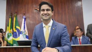 Vereador Pedro Matos sorrindo durante a cerimônia de posse na Câmara Municipal de Fortaleza, com bandeiras ao fundo.