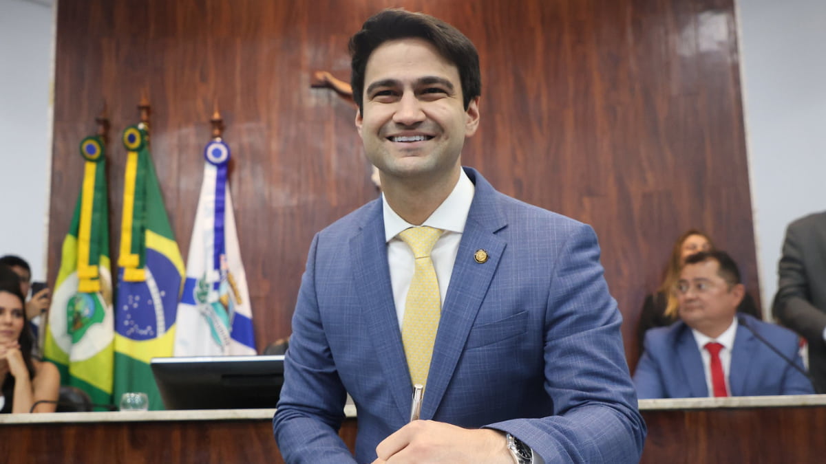Vereador Pedro Matos sorrindo durante a cerimônia de posse na Câmara Municipal de Fortaleza, com bandeiras ao fundo.
