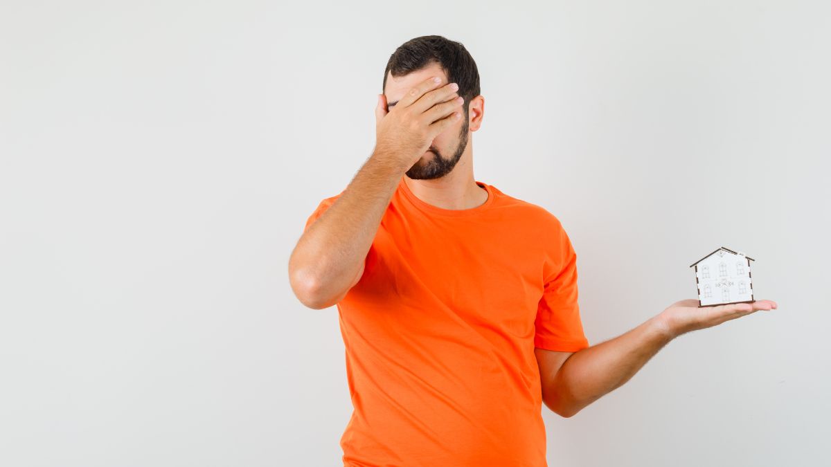Homem de camisa laranja tapando os olhos enquanto segura uma casa em miniatura, simbolizando incertezas diante dos preços de imóveis.