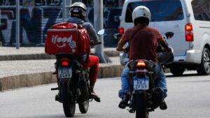 Dois motociclistas em uma rua, um deles entregador com mochila do iFood, representando o aumento das vendas de motos no Brasil, impulsionado pelo crescimento de serviços de entrega.