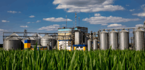 A imagem simboliza o agronegócio, mostrando uma instalação industrial com grandes silos e uma plantação