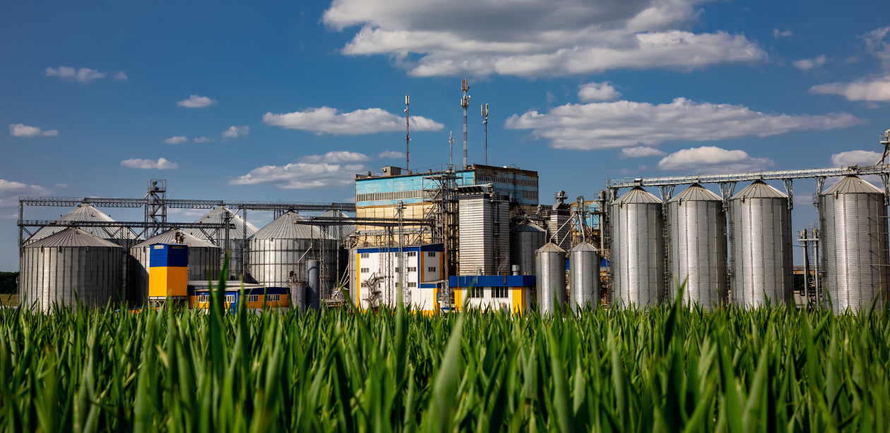 A imagem simboliza o agronegócio, mostrando uma instalação industrial com grandes silos e uma plantação