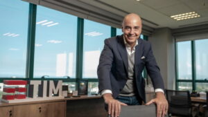 Alberto Griselli, CEO da TIM, sorrindo em seu escritório com vista panorâmica, ao lado de uma placa da TIM.