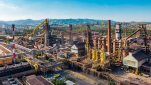 Vista aérea de uma siderúrgica com diversas estruturas industriais, incluindo altos-fornos, tubulações e chaminés, cercada por montanhas e áreas urbanas ao fundo. Representação da Usiminas, envolvida na disputa judicial sobre a venda de ações da CSN.