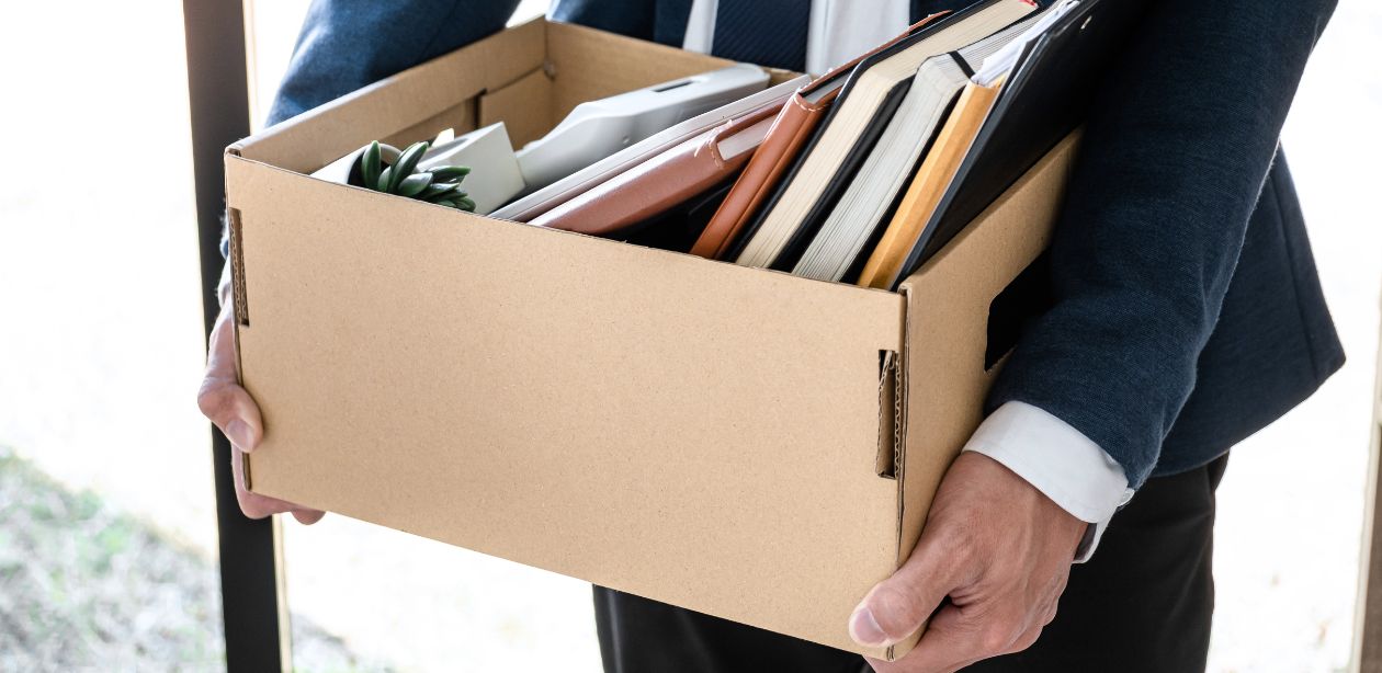 A imagem mostra um homem de terno segurando uma caixa de papelão com itens pessoais de escritório, representando as carreiras em risco pela popularização da IA.