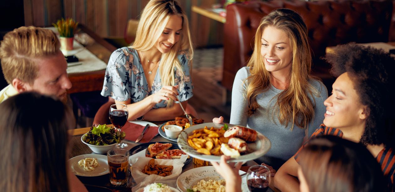 A imagem mostra um grupo de amigos em volta de uma mesa de restaurante, com a gastronomia de experiência.