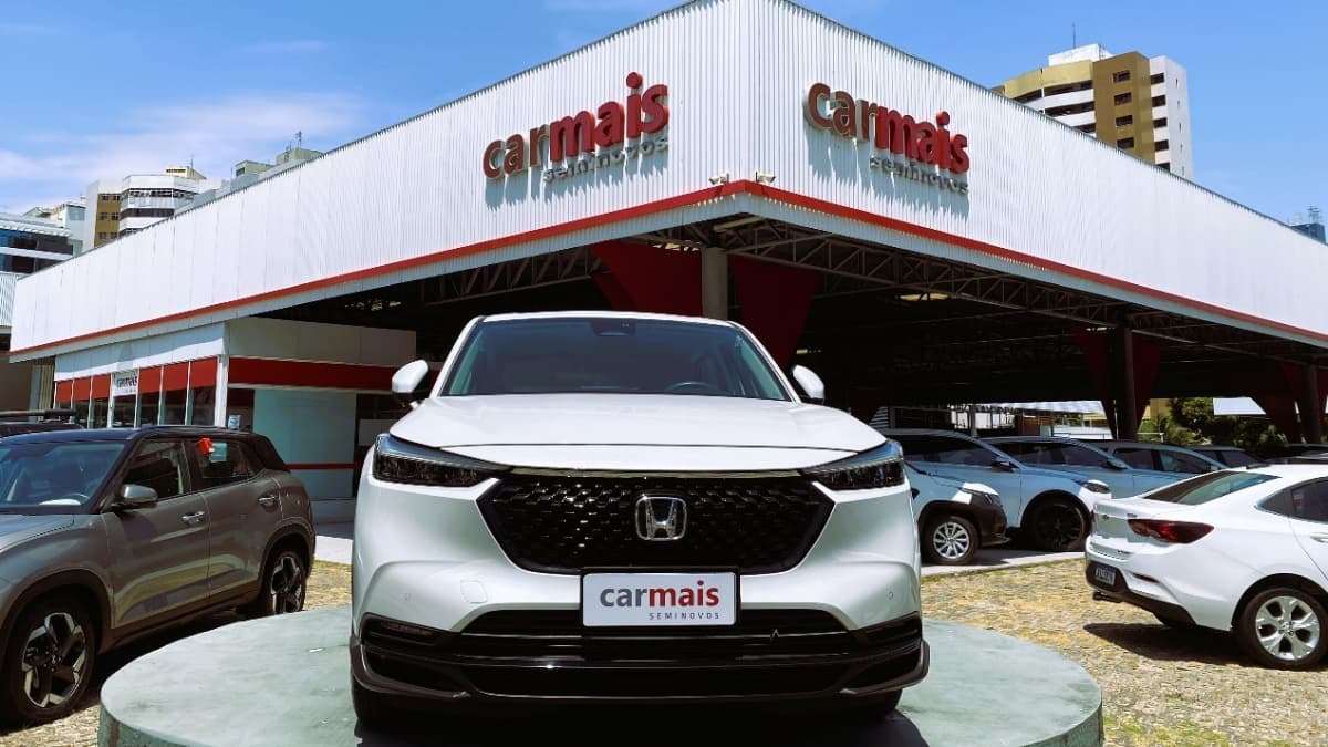 Fachada da concessionária Carmais Seminovos com veículos em exposição, destacando um Honda branco na frente, sob um céu azul.