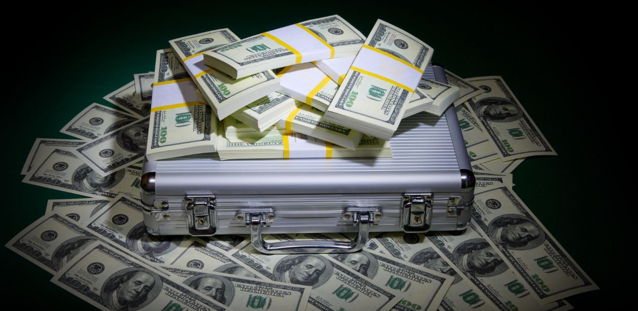 A imagem mostra uma maleta e várias notas de dólar em volta, simbolizando a cotação do dólar.