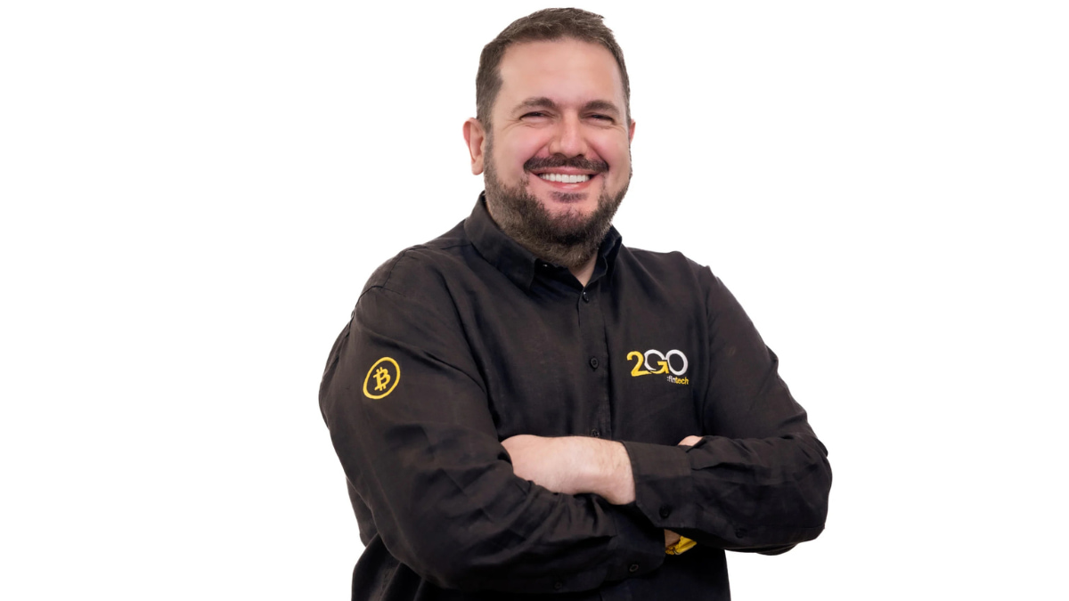 Cyllas Elia, CEO da 2GO Bank, sorridente e de braços cruzados, vestindo camisa preta com logotipo da fintech investigada por lavagem de dinheiro e símbolo de Bitcoin na manga.