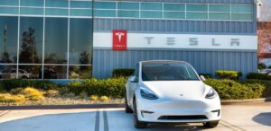 A imagem mostra um carro da Tesla em frente a facha de uma das unidades da empresa, representando a queda de vendas da Tesla.