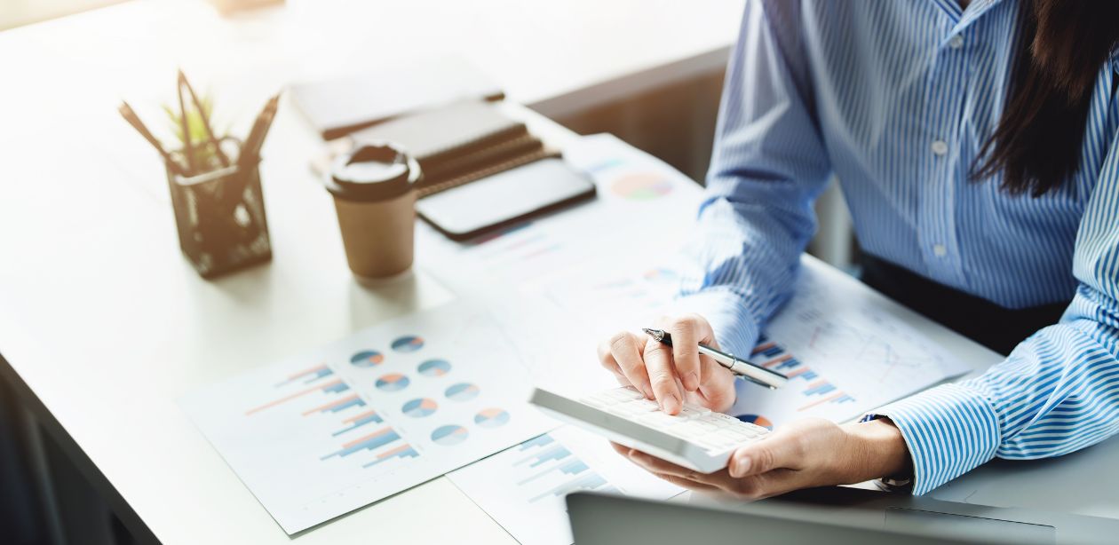 A imagem mostra uma pessoa sentada em uma mesa, fazendo o cálculo com uma calculadora. Na mesa há papéis, cadernos, copo de café e vários itens de escritório, mostrando como a organização das finanças para autônomos é importante.
