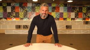 Gustavo de Lima Furtado, presidente do Grupo SBF, posa sorridente apoiado em uma mesa, com um fundo colorido e moderno, representando a identidade da empresa.