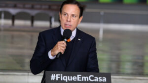 oão Doria discursa segurando um microfone ao lado de um púlpito com a mensagem "#FiqueEmCasa". A imagem está relacionada à ButanVac, vacina contra a Covid desenvolvida pelo Instituto Butantan.