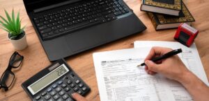 A imagem mostra uma mesa, acima dela um notebook aberto, uma pessoa fazendo cálculos em uma calculadora e fazendo anotações em um papel, simbolizando a declaração do Imposto de Renda 2025.