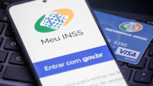 A imagem mostra um celular e na tela aparece o início do app Meu INSS, ao fundo aparece um cartão de benefício para representar o pagamento INSS que acontece nessa segunda.