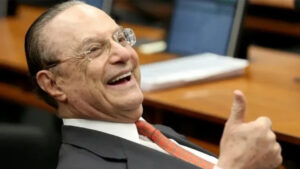 Paulo Maluf sorrindo e fazendo um gesto de positivo com o polegar, usando terno e gravata, em um ambiente formal com mesas e notebooks ao fundo.