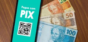 A imagem mostra um celular com um QR Code para pagamento via Pix e ao lado, notas de dinheiro, simbolizando a tecnologia do Pix na economia.