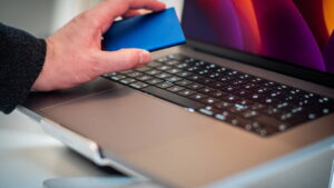 Mão segurando um cartão azul próximo ao teclado de um notebook moderno, simbolizando compras online e pagamentos digitais.