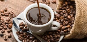 A imagem mostra uma xícara de café cheia, com grãos de café em volta, representando os alimentos afetados pela inflação.