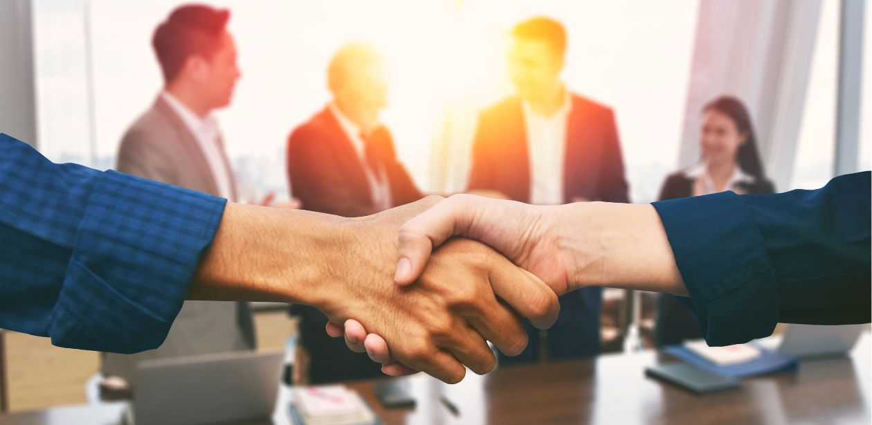 A imagem mostra empresários desfocados ao fundo e duas pessoas apertando as mãos à frente da imagem.