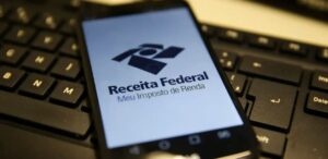 A imagem mostra um celular com a tela acesa com a logo da Receita Federal na tela, representando a restituição do Imposto de Renda.