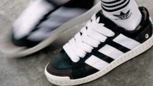 Tênis Adidas preto e branco com listras clássicas e cadarços largos, usado com meia listrada da marca, em movimento sobre o chão.