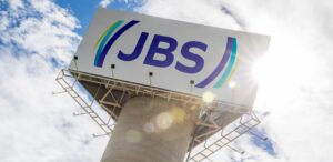 A imagem mostra o logo da JBS em um painel externo para representar que as ações da JBS disparam com acordo e podem valorizar até 98%.
