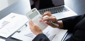 A imagem mostra uma pessoa fazendo cálculos em uma calculadora e vários papéis na mesa para representar a carga tributária.