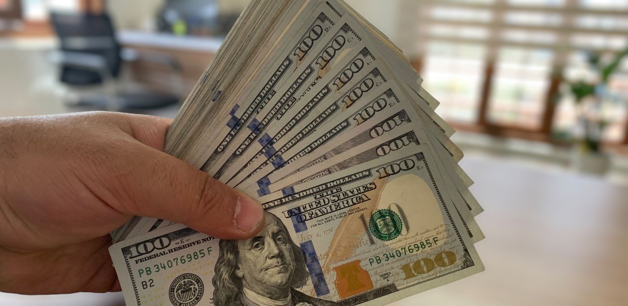 A imagem mostra diversas notas de dólares para representar o mercado financeiro.