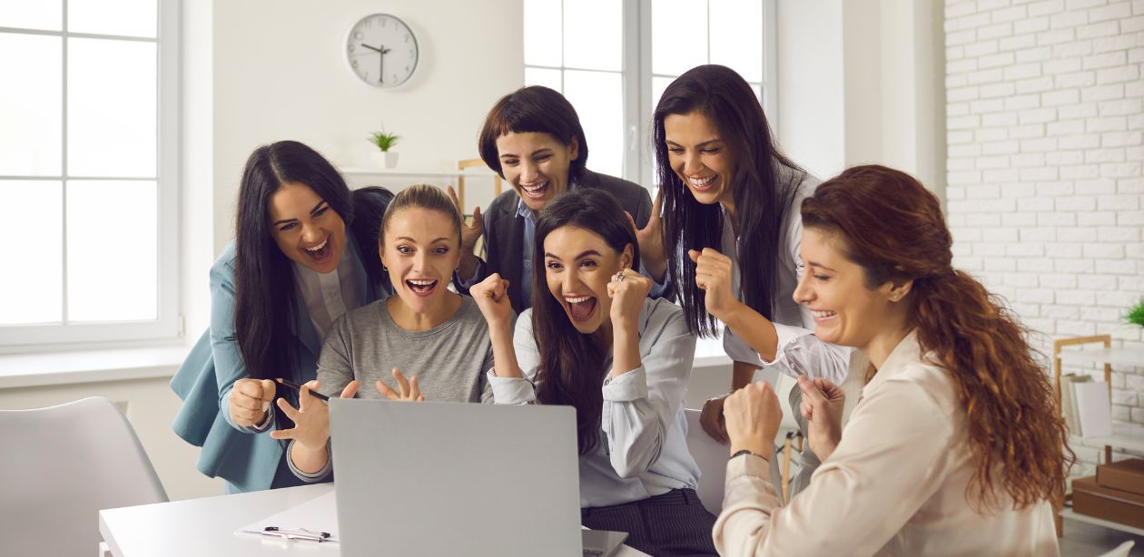 A imagem mostra seis mulheres em volta de um notebook comemorando para simbolizar as mulheres investidoras na B3.