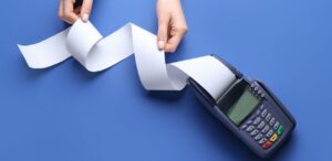 A imagem mostra uma maquinha de cartão e uma pessoa segurando o papel da maquininha.