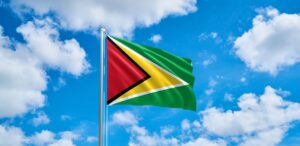 A imagem mostra a bandeira da Guiana e ao fundo, o céu para representar o PIB per capita da Guiana.