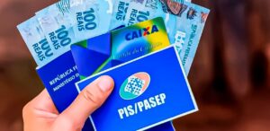 A imagem mostra uma pessoa segurando o cartão de saque do PIS/PASEP e notas de dinheiro.